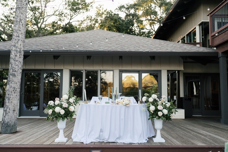 Sweetheart table on the venue deck framed by large white floral urns and string lights against the building facade.