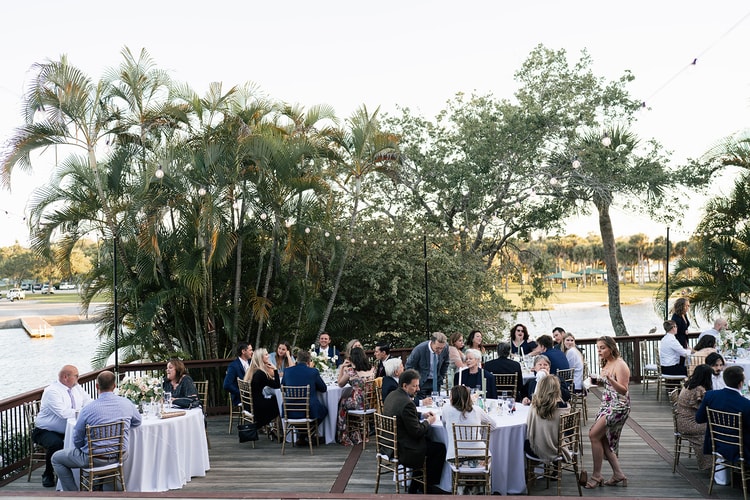 Reception dinner in full swing on a waterfront deck at dusk, guests dining under palm trees with the bay visible beyond.