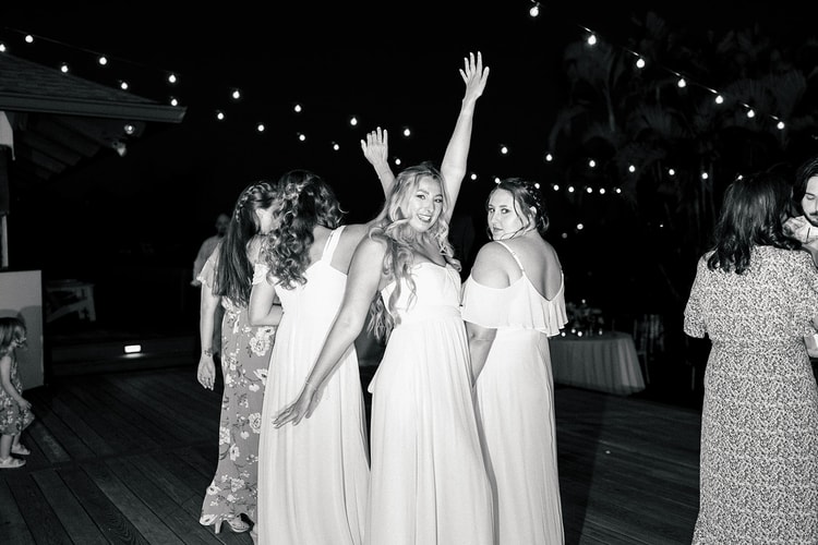 Bridesmaids dancing on the outdoor reception deck at night, string lights overhead, one with her arm raised in celebration.