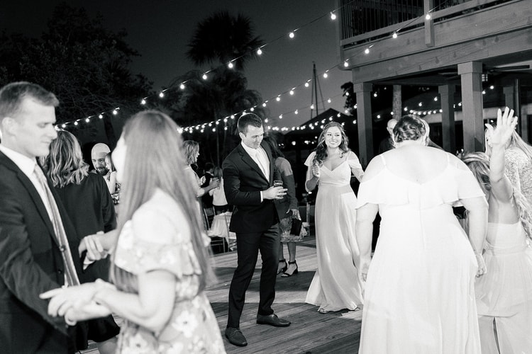 Guests mingling and dancing at the outdoor waterfront reception, lit by string lights against a darkening sky.