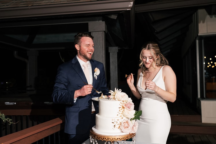 Bride and groom laughing together at the cake table after cutting the wedding cake, the decorated two-tier cake before them.