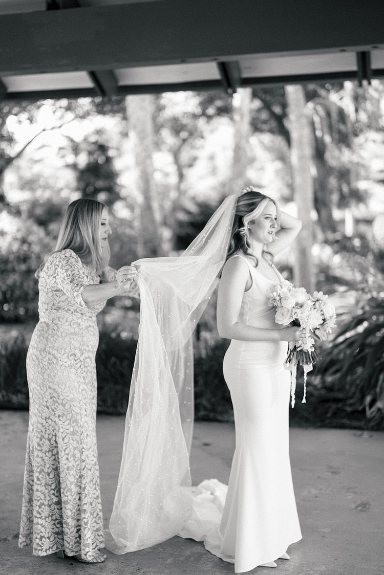 Black-and-white image of a family member helping the bride fluff her cathedral veil just before the ceremony processional.