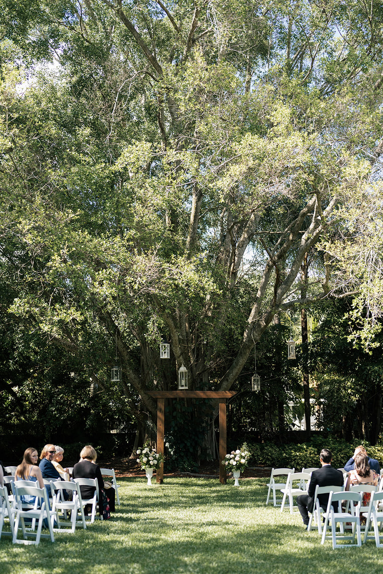 Outdoor ceremony setup: rows of white chairs face a wooden arch hung with lanterns beneath a towering oak tree.