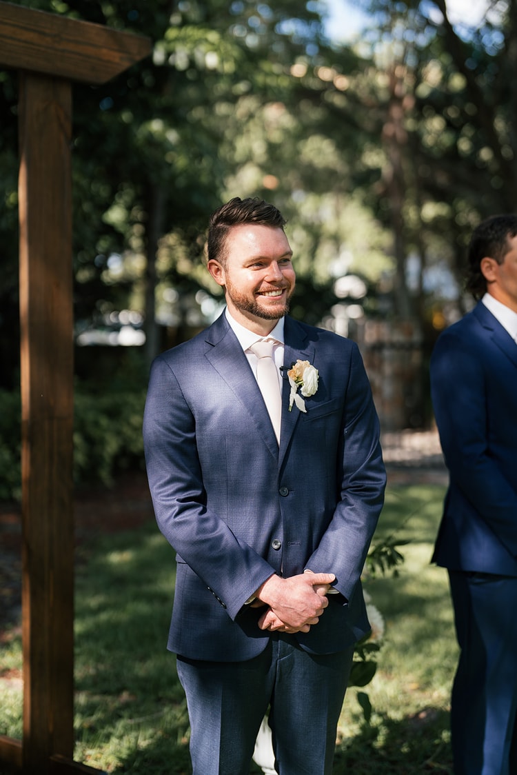 Groom beaming at the altar beside a wooden arch as the bride approaches down the aisle.