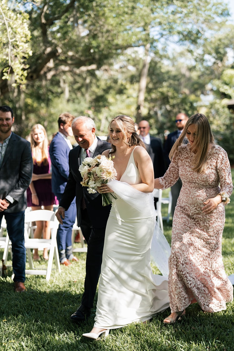 Bride walks the aisle escorted by both parents, all three smiling as guests stand and look on in the garden setting.