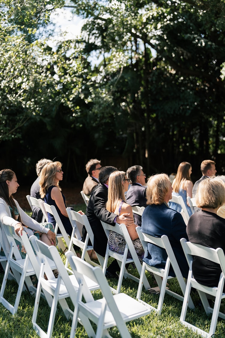 Ceremony guests seated in white folding chairs outdoors, watching attentively in dappled sunlight.