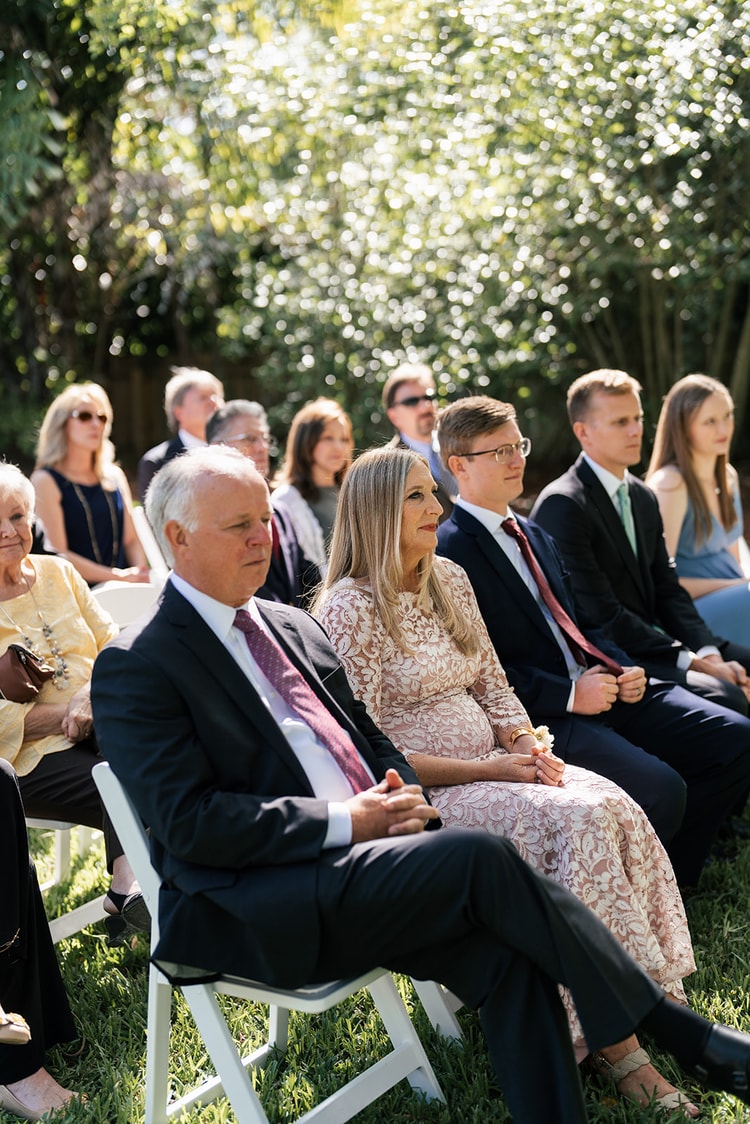 Ceremony guests seated in white chairs on the sunlit lawn, watching the proceedings with focused attention.
