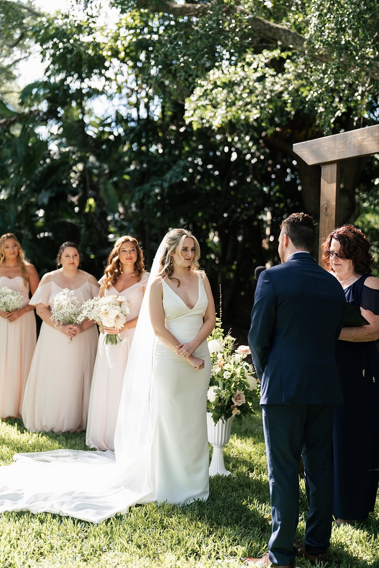 Bride standing at the altar with bridesmaids in blush behind her, groom facing her as the officiant reads from her notes.