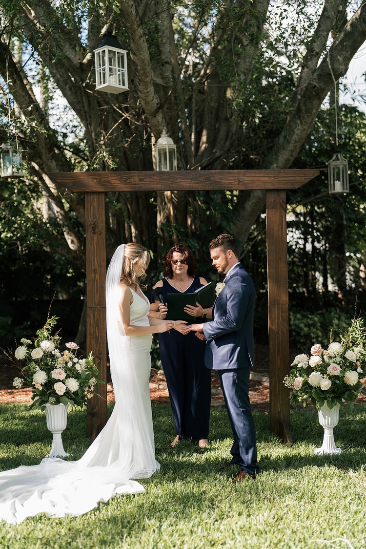 Bride and groom exchange vows under a wooden arch adorned with white floral urns, officiant beside them.