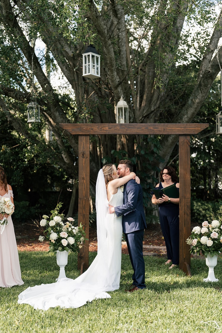 Newlyweds share their first kiss under the wooden ceremony arch flanked by white flower arrangements.