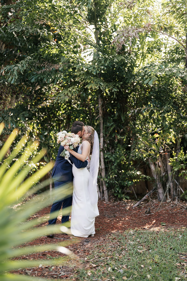 Bride and groom kissing in a shaded garden path framed by tropical palms and ferns.