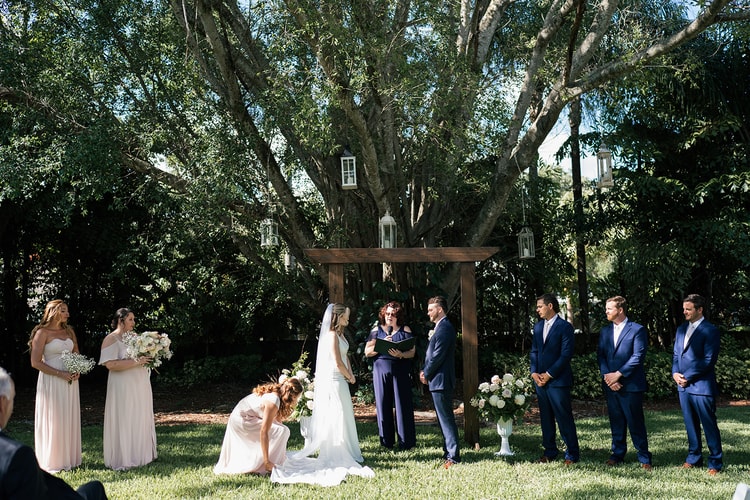 Ceremony moment: a bridesmaid kneels to arrange the bride's train at the altar while the couple stands before the officiant.