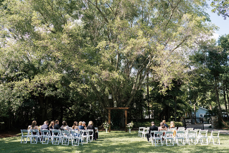 Wide shot of the outdoor ceremony lawn with guests seated in white chairs under a canopy of trees, wooden arch in the background.