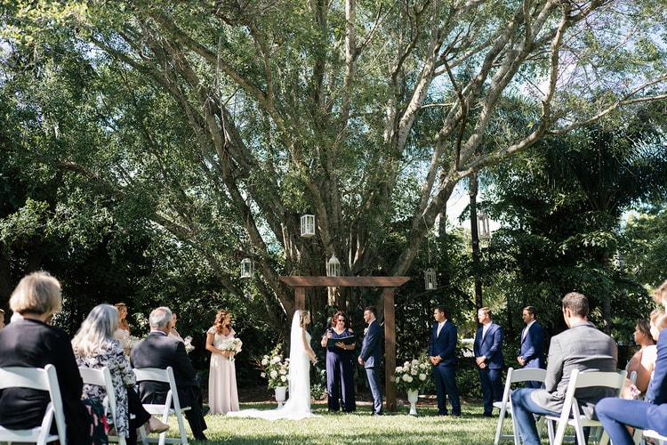 Wide ceremony scene with couple at the wooden arch under tall trees, bridal party and guests flanking both sides.