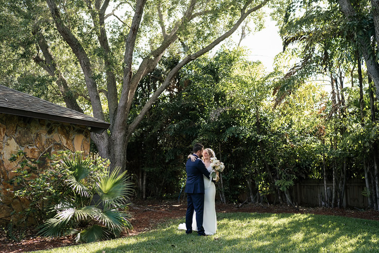 Couple embracing in a secluded garden corner, large leafy tree and stone wall backdrop.