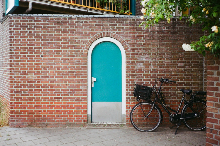 Teal arched door set into a red brick wall beside a bicycle with a crate basket and a flowering rosebush.