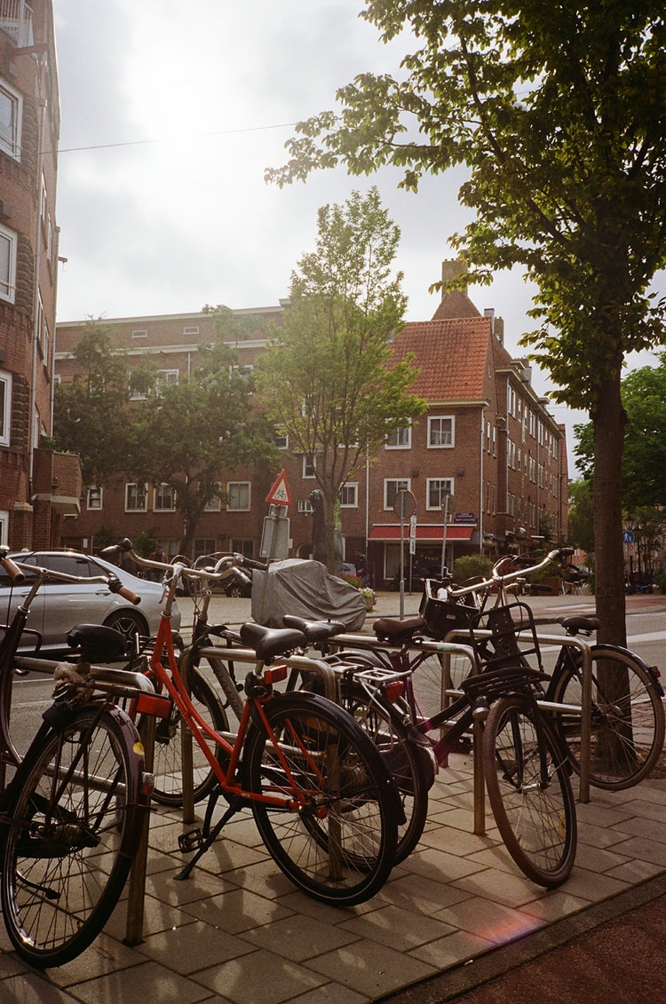 Rows of Dutch bicycles parked at a curbside rack on an Amsterdam street corner with brick apartment buildings behind.