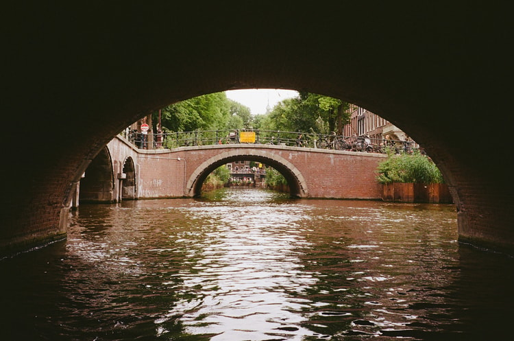 View from a canal boat passing beneath a stone bridge in Amsterdam, with a second arched bridge visible ahead.