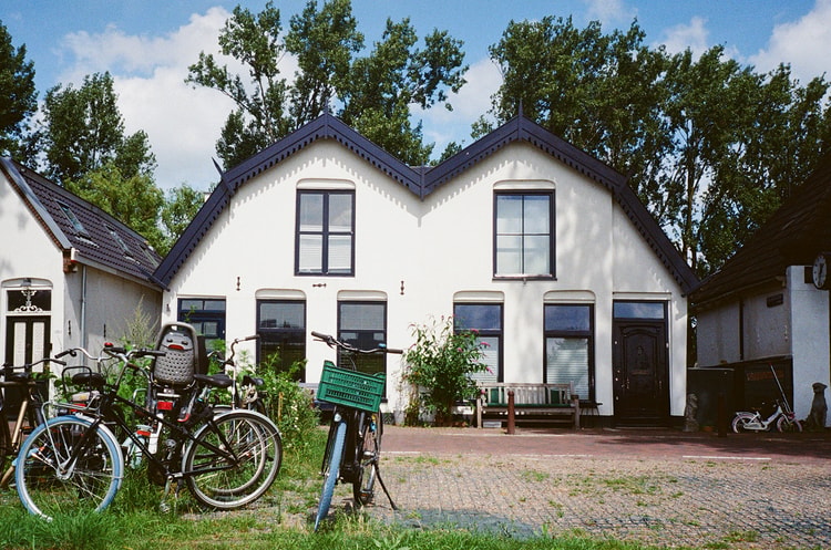 White Dutch cottage with a distinctive double-peaked navy roof set among bicycles on brick pavement.