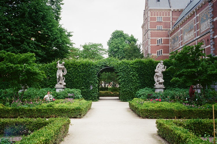 Classical statues flank a hedge-lined garden path leading to an archway beside the Rijksmuseum in Amsterdam.