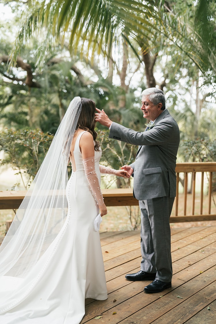 Bride's father lifts her veil on a wooden deck surrounded by lush tropical trees during a first look, wedding photography
