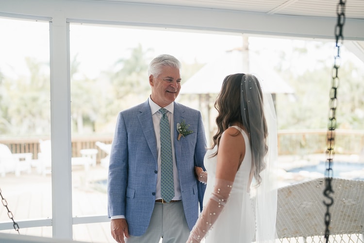 Father in a blue blazer smiles warmly at the bride in her pearl-sleeved gown on a bright screened porch, wedding photography