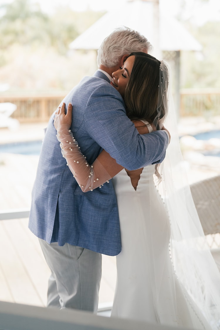 Bride in a pearl veil embraces her father in a light blue jacket on a sunlit porch, both smiling, wedding photography