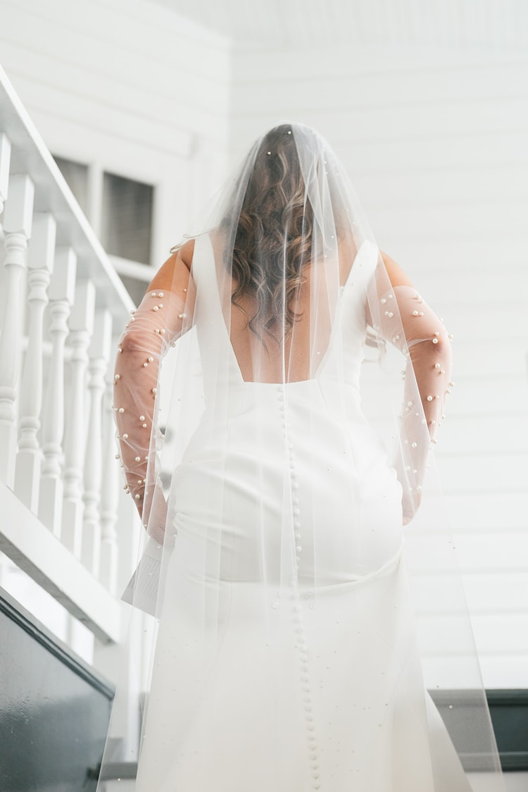 Bride shot from behind on a white staircase, pearl veil and sheer pearl-dotted gloves cascading down her open-back gown, wedding photography