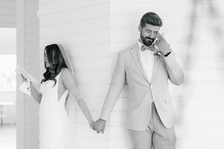 Black and white: groom in a tan suit wipes a tear while holding the bride's hand around a white-painted column, vow books in hand, wedding photography