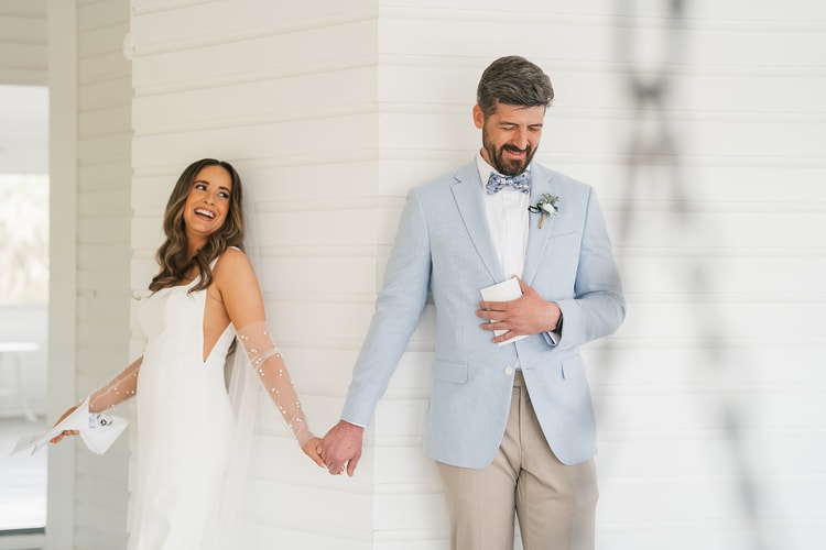 Bride beams over her shoulder at the groom as they hold hands around a column, both holding their vow books, wedding photography