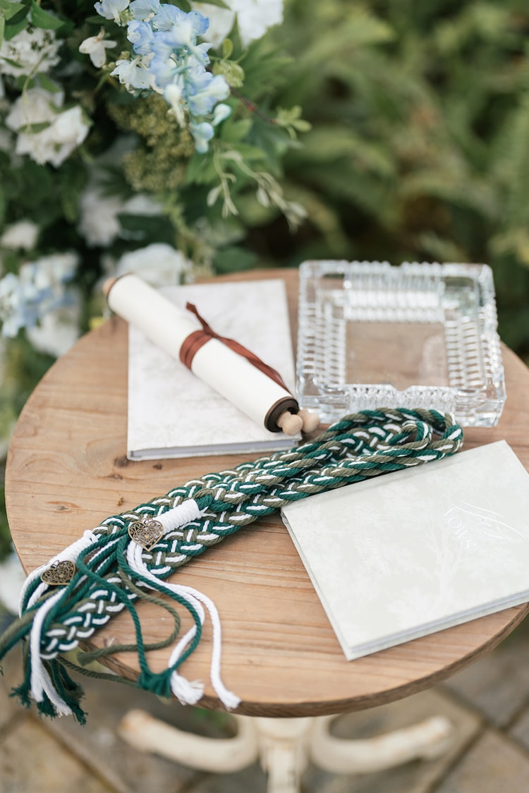 Ceremony table detail: braided green and white handfasting cord, vow scroll, crystal ring dish and open book beside blue and white flowers, wedding details