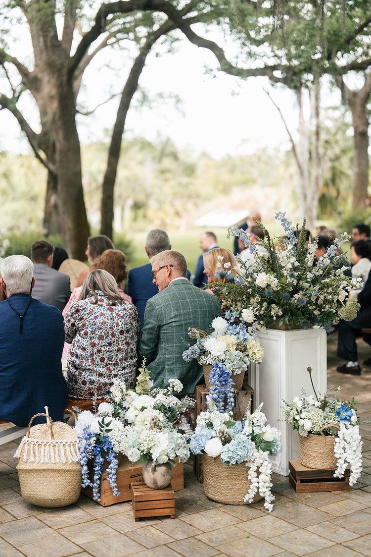 Guests seated at an outdoor ceremony viewed from behind, blue and white floral arrangements and wicker baskets lining the brick aisle, wedding photography
