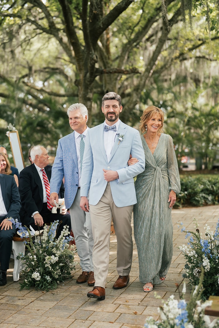 Groom in a light blue suit walks the aisle flanked by his parents under sweeping moss-draped oaks, smiling broadly, wedding photography