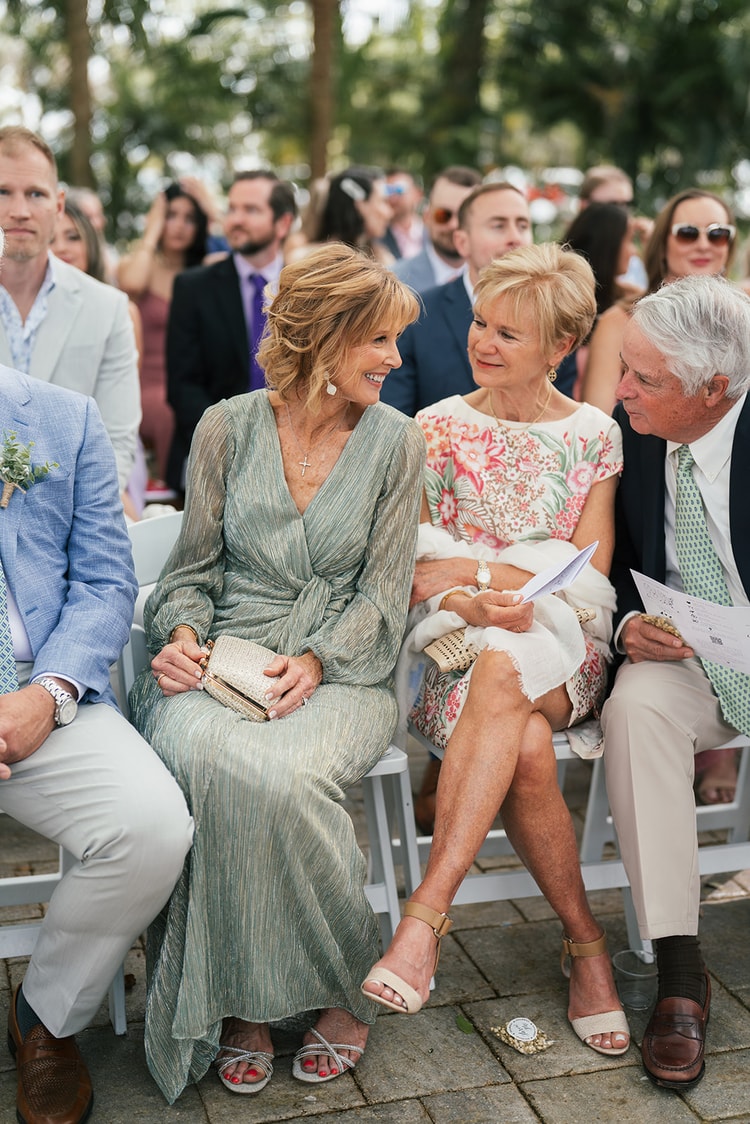 Guests chat in their seats at an outdoor ceremony, women in sage and floral dresses enjoying the moment before the procession, wedding photography