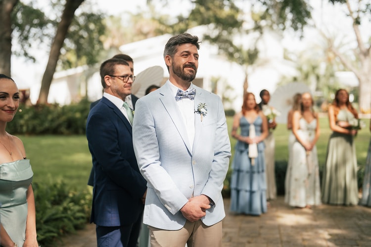 Groom in a light blue suit and floral bow tie stands at the altar watching the processional, bridesmaids in sage and blue behind him, wedding photography