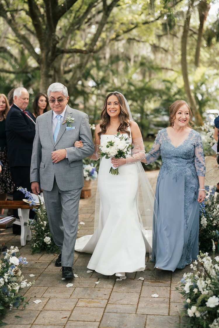 Bride smiles radiantly walking the aisle between her parents, white bouquet in hand, blue floral arrangements lining the brick path, wedding photography