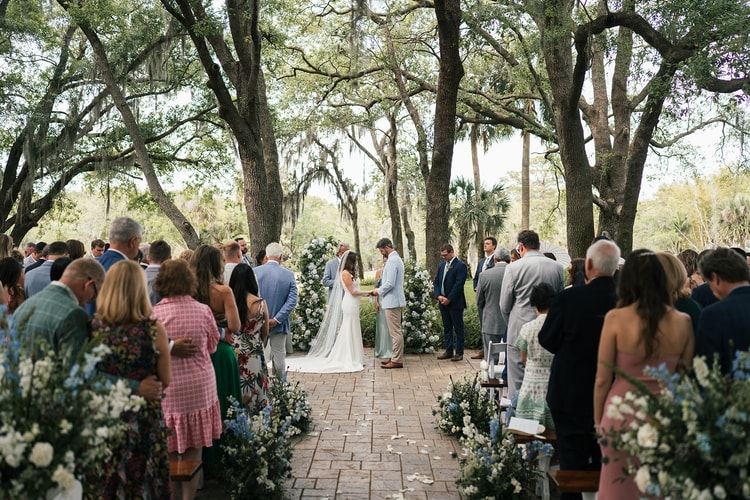 Wide shot of the couple at the altar beneath tall oaks, guests standing and watching from both sides of the brick aisle, wedding photography