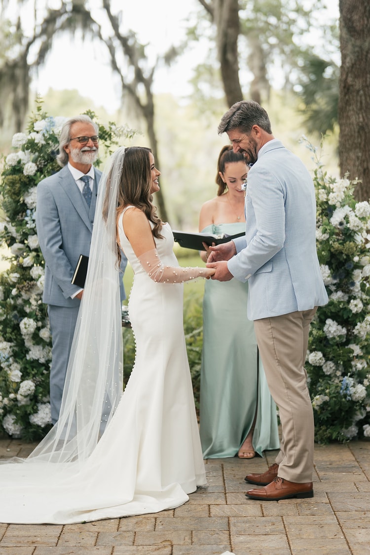 Bride and groom exchange rings at the altar before a lush white and blue floral arch, officiant and friends close by, wedding photography