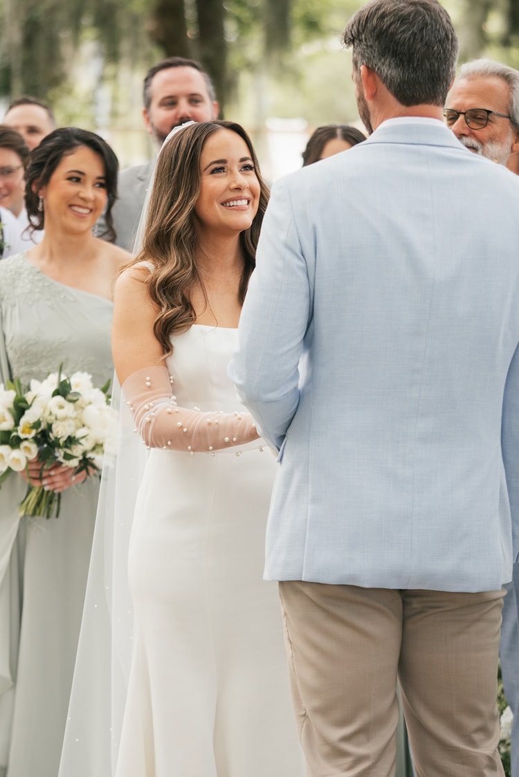 Bride gazes up at the groom with a radiant smile, bridesmaids in sage gowns holding white bouquets visible behind her, wedding photography