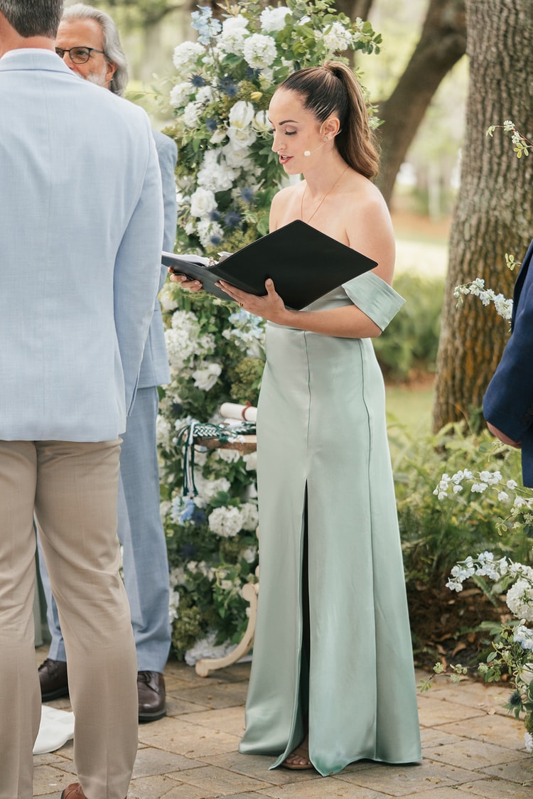 Officiant in a sage off-shoulder gown reads from a black folio before the white and blue floral ceremony arch, wedding photography