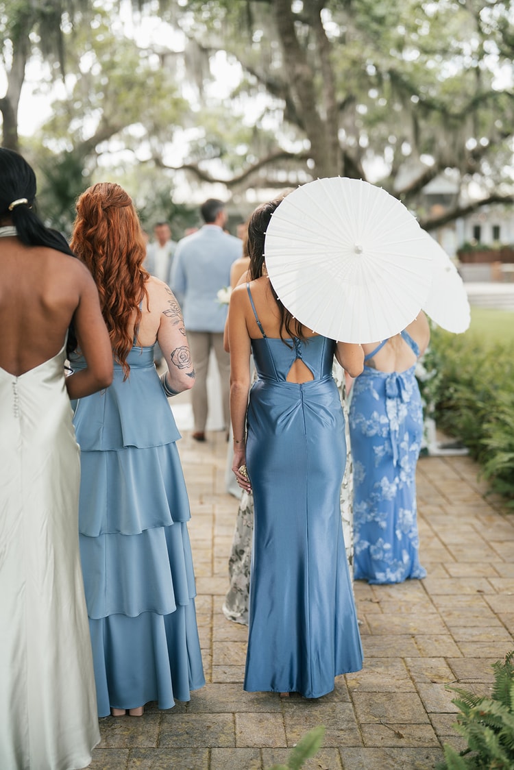 Bridesmaids in blue and floral gowns walk away from camera along a brick path beneath moss-draped oaks, one carrying a white parasol, wedding photography
