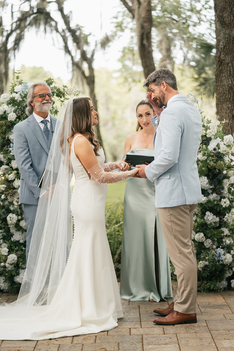Couple exchange vows at the altar before a lush blue and white floral arch, officiant smiling between them, wedding photography