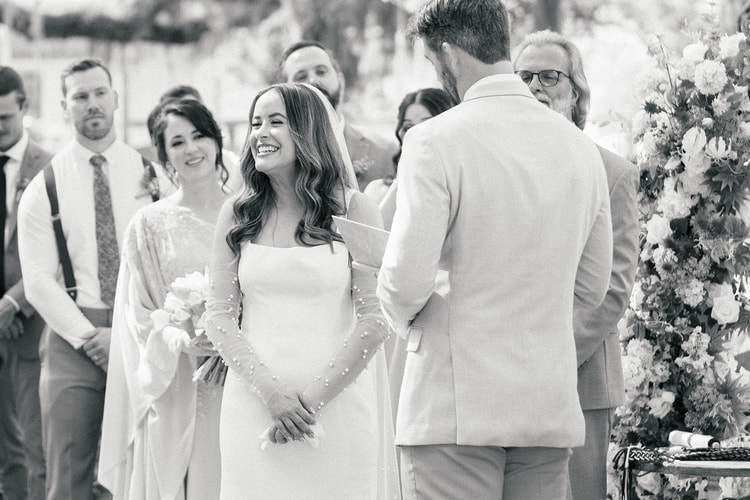 Black and white: bride laughs joyfully as the groom reads his vows beside the ceremony arch, guests visible in soft focus behind, wedding photography