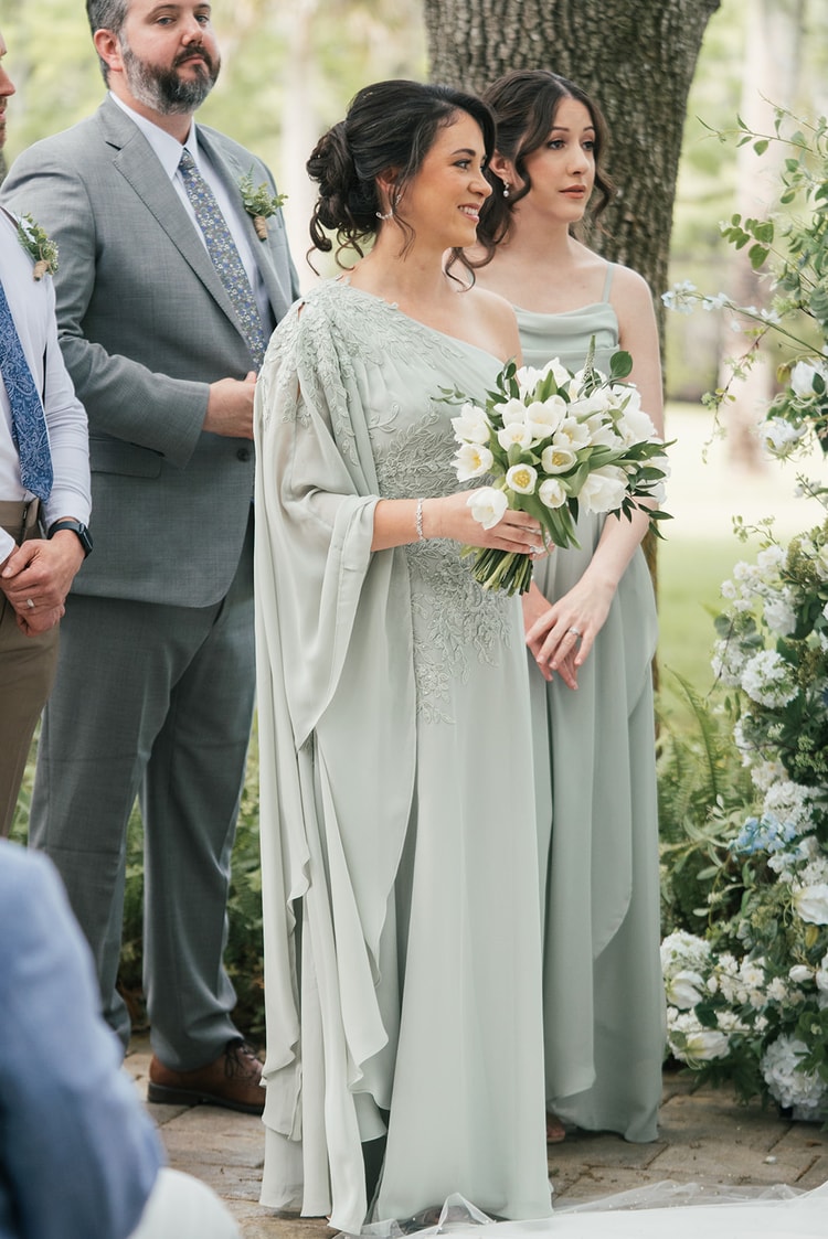 A woman in an embroidered sage cape gown holds a white tulip bouquet and smiles at the altar beside another attendant, wedding photography