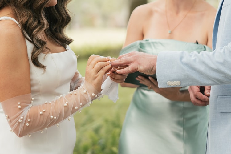 Close-up of the bride placing a ring on the groom's finger, her pearl-beaded sleeve visible, a bridesmaid in sage looking on.