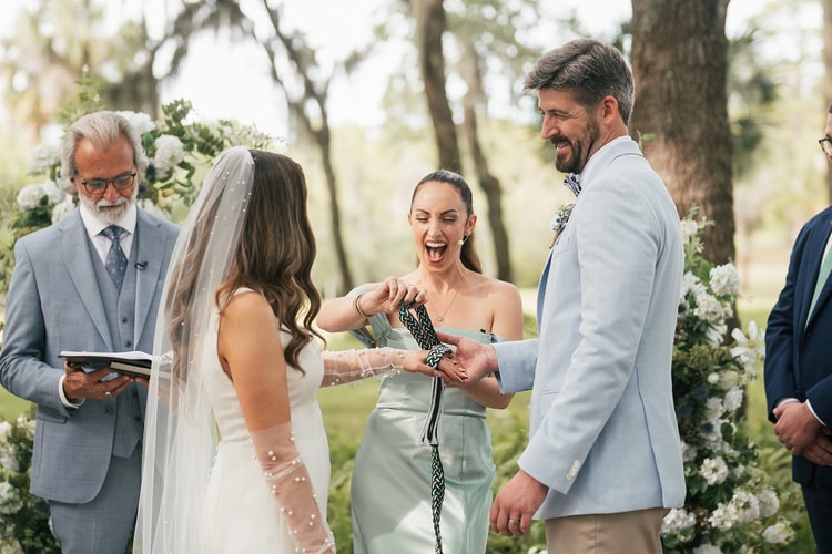 Officiant laughs with delight while wrapping a braided green cord around the couple's joined hands during a handfasting ritual, wedding photography