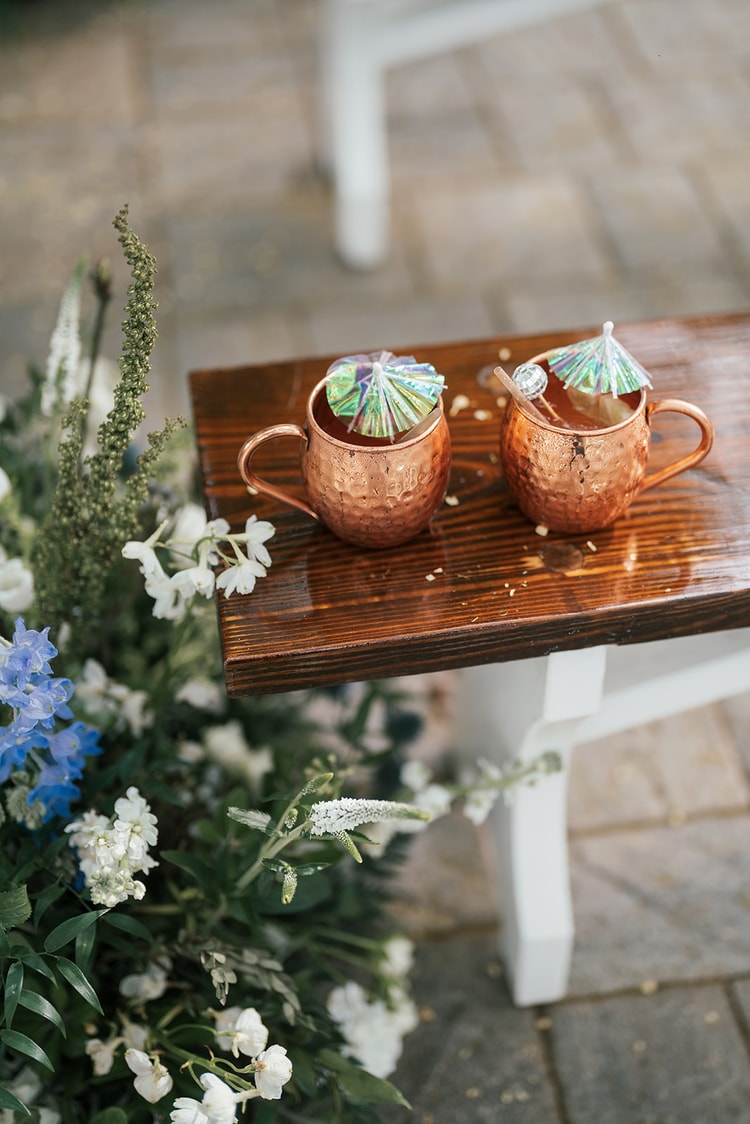 Two copper mule mugs with paper umbrellas sit on a wooden bench beside blue delphinium and white florals at the reception.
