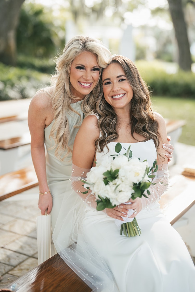Bride sits holding a white peony bouquet as a bridesmaid in sage leans in cheek-to-cheek for a joyful portrait, wedding photography