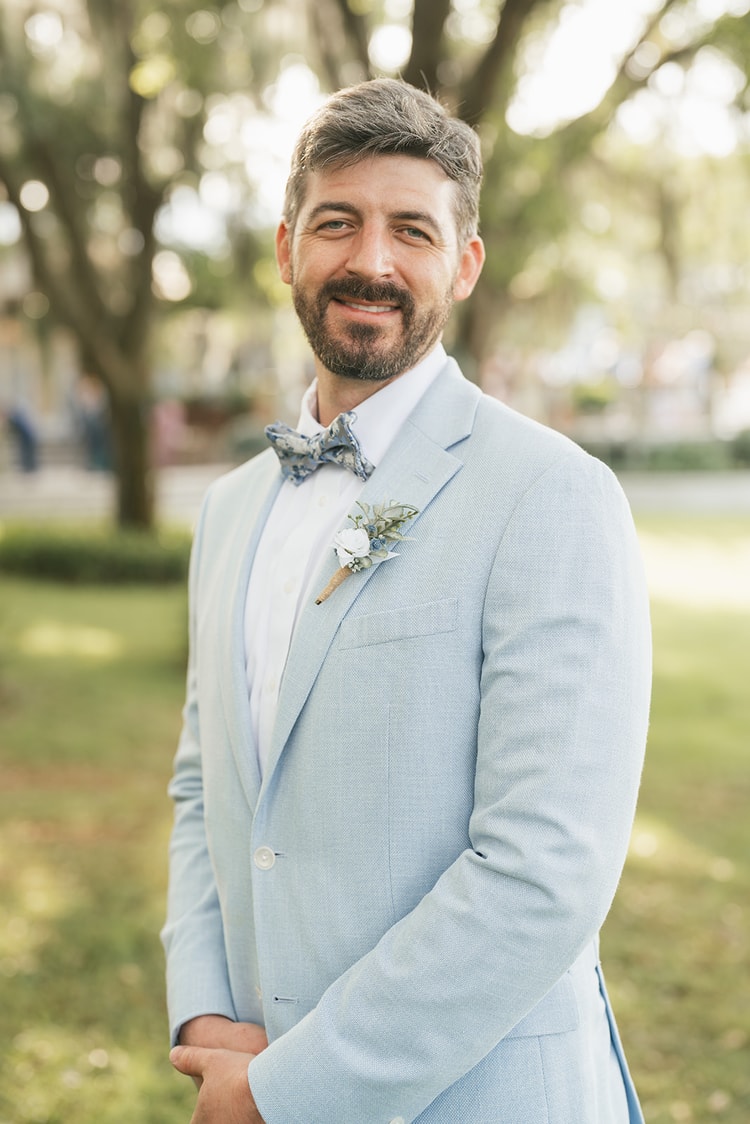 Groom portrait: bearded groom in a light blue suit and floral bow tie with a white boutonniere, smiling against soft bokeh greenery, wedding photography