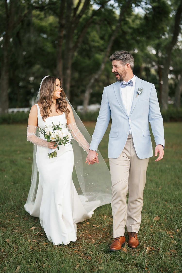 Couple stroll hand in hand on a lawn, bride glancing at the groom with a smile, her pearl veil billowing, tall trees behind them, wedding photography